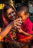 Sayra Begum (m.), profitiert von ihrer Regenwassergewinnung, die von CCDB gestellt wurde und kann jetzt sauberes Trinkwasser aus dem Wassertank zapfen und ihrem Sohn geben. Die Menschen in den kleinen Siedlungen an den Fluessen in der Naehe von Shyamnagar in Sued Bangladesch, spuehren deutlich den Klimawandel. Der Meeresspiegel steigt und Daemme werden zerstört. Da durch werden Felder und die wichtigen Suesswasserteiche ueberflutet und versalzen. CCDB Bangladesch arbeitet an Suesswassergewinnungs-Projekten und sorgt dafuer, dass die Menschen nach und nach sauberes Trinkwasser erhalten. Fotoserie Bangladesch